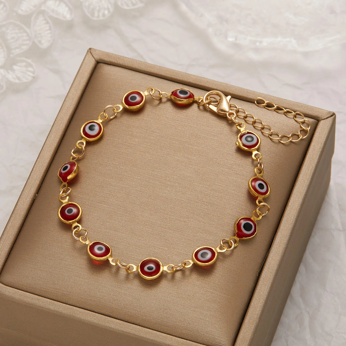 Gold charm bracelet with red and white evil eye beads displayed in a light brown box.