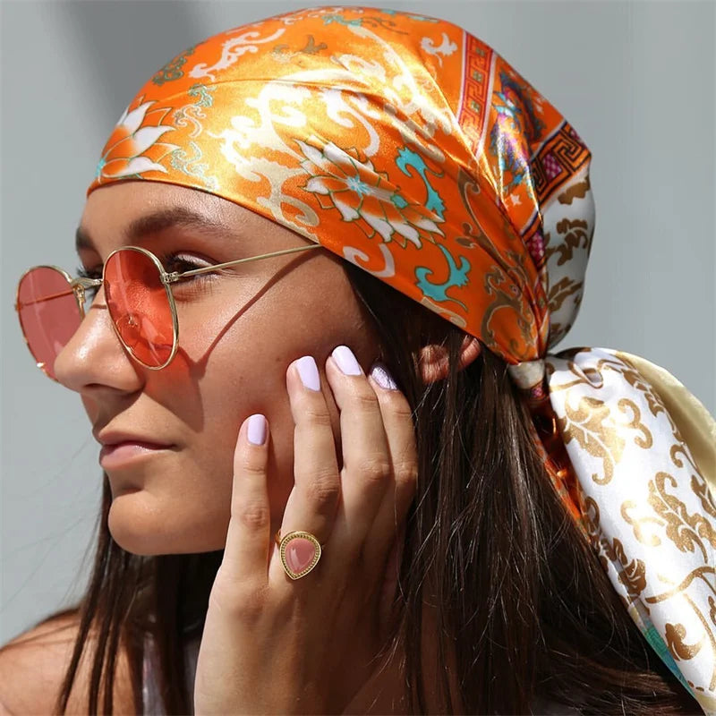 A woman wearing an orange satin square scarf tied around her head and hair, paired with round sunglasses.