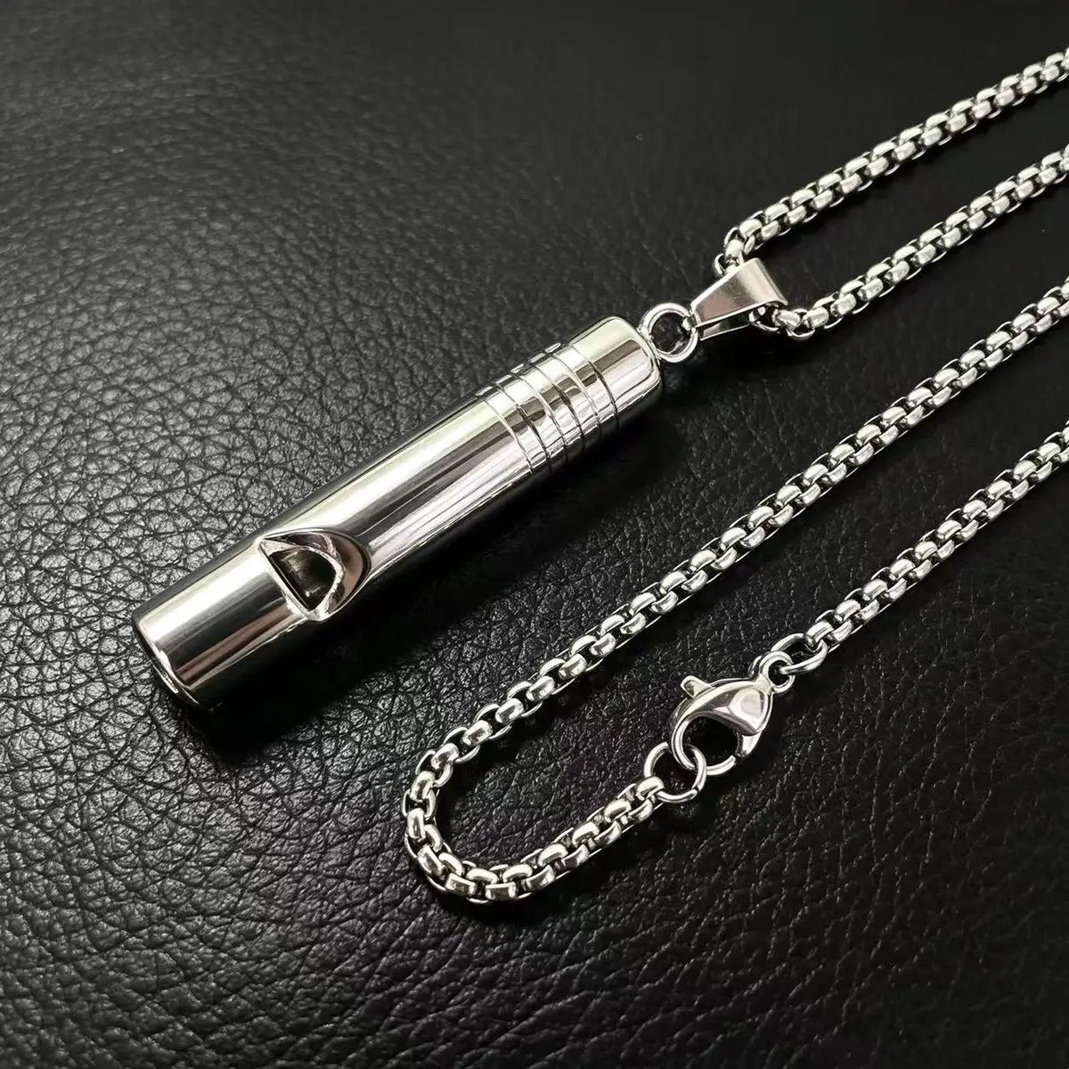 A silver stainless steel whistle pendant necklace for men displayed on a black leather surface.