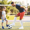 A young man in a dark shirt and orange shorts is practicing soccer with a young boy on a field.