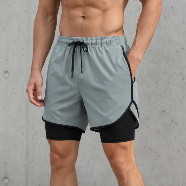 Un hombre vistiendo shorts deportivos grises de secado rápido con ribetes negros y cordón, de pie contra un fondo de concreto