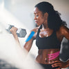 A woman in a black sports bra is performing an exercise while applying professional kinesiology tape to her shoulder.