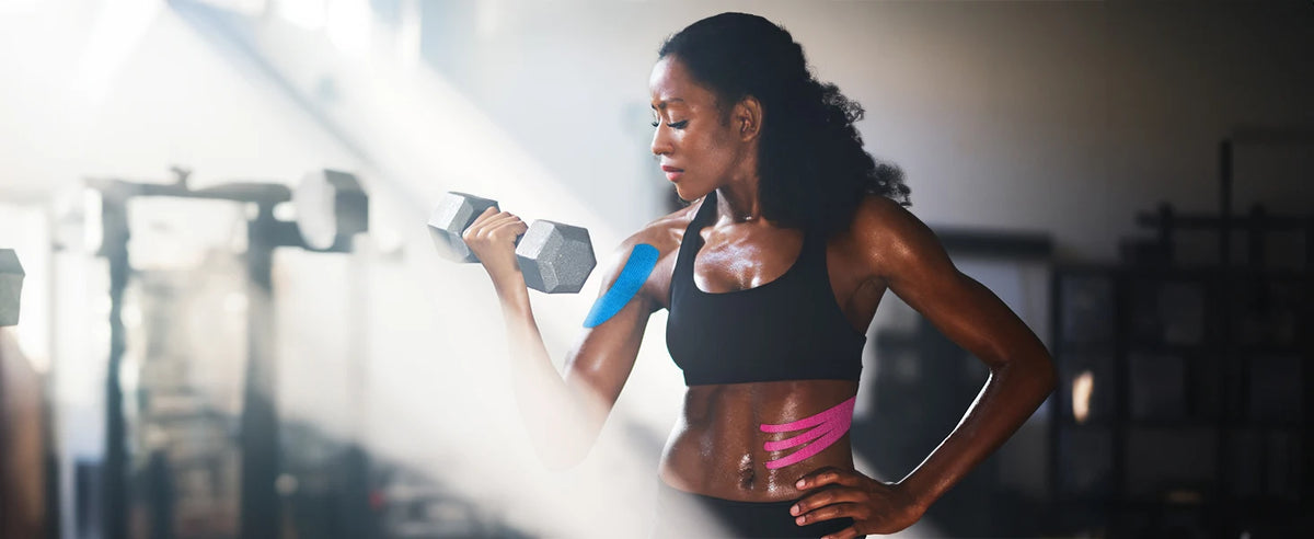 A woman in a black sports bra is performing an exercise while applying professional kinesiology tape to her shoulder.