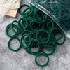 A pile of dark green elastic hair bands is shown on a gray surface, with a clear plastic container partially visible.