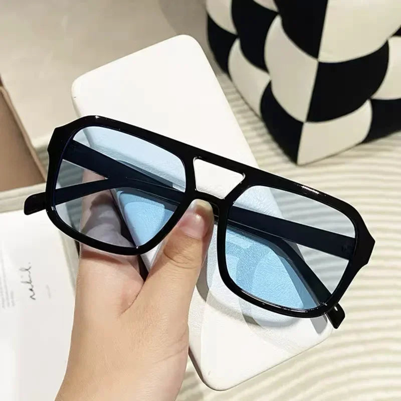 Un par de gafas de sol cuadradas con montura negra y lentes azul claro, sobre una superficie blanca.
