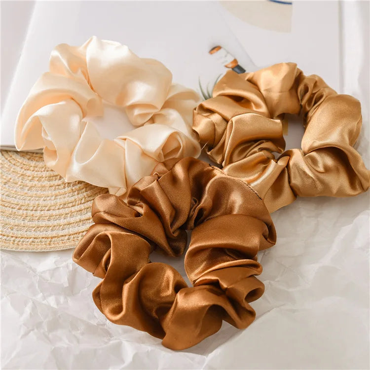 A set of satin scrunchies in shades of beige and gold are displayed on white paper.
