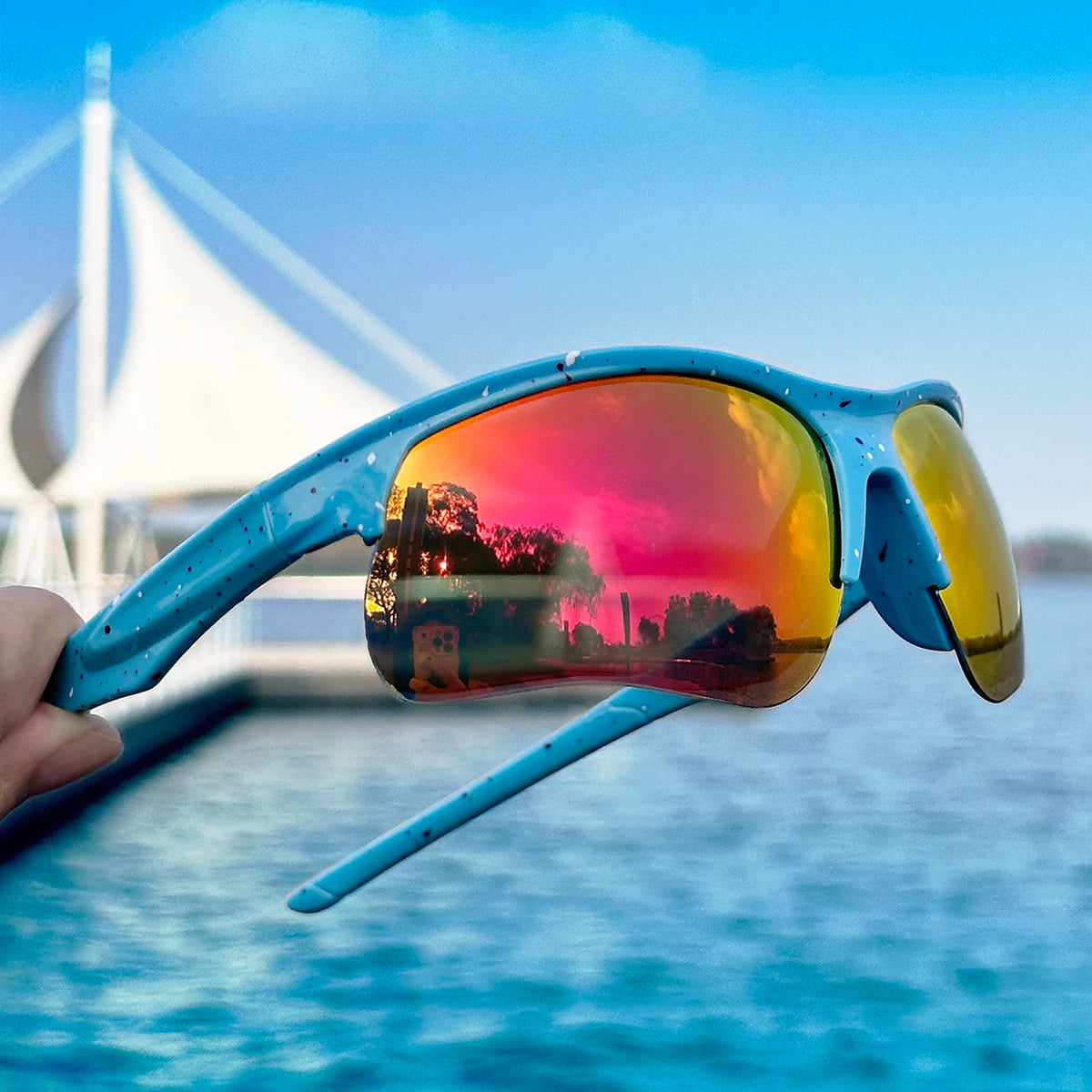 A close-up of blue sports sunglasses with polarized lenses reflecting a sunset over water and sailboats.