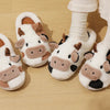 A pair of cartoon cow winter home slippers in white and brown fur are shown on a wooden floor.