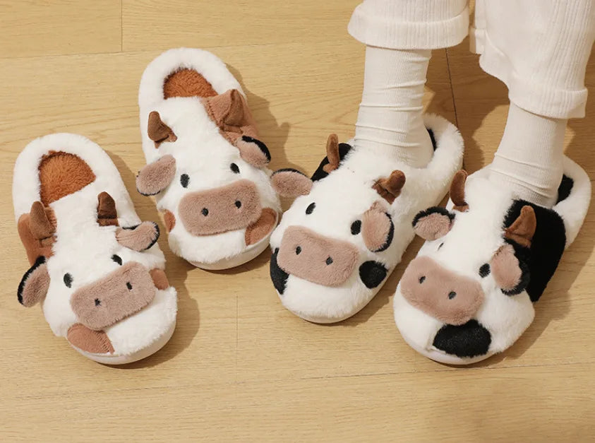 A pair of cartoon cow winter home slippers in white and brown fur are shown on a wooden floor.