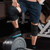 A person wearing dark athletic shorts and knee wraps is performing a strength training exercise with a weight plate.