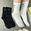 A pair of white crew socks with black crosses and a pair of black crew socks with silver crosses are displayed on a gray surf