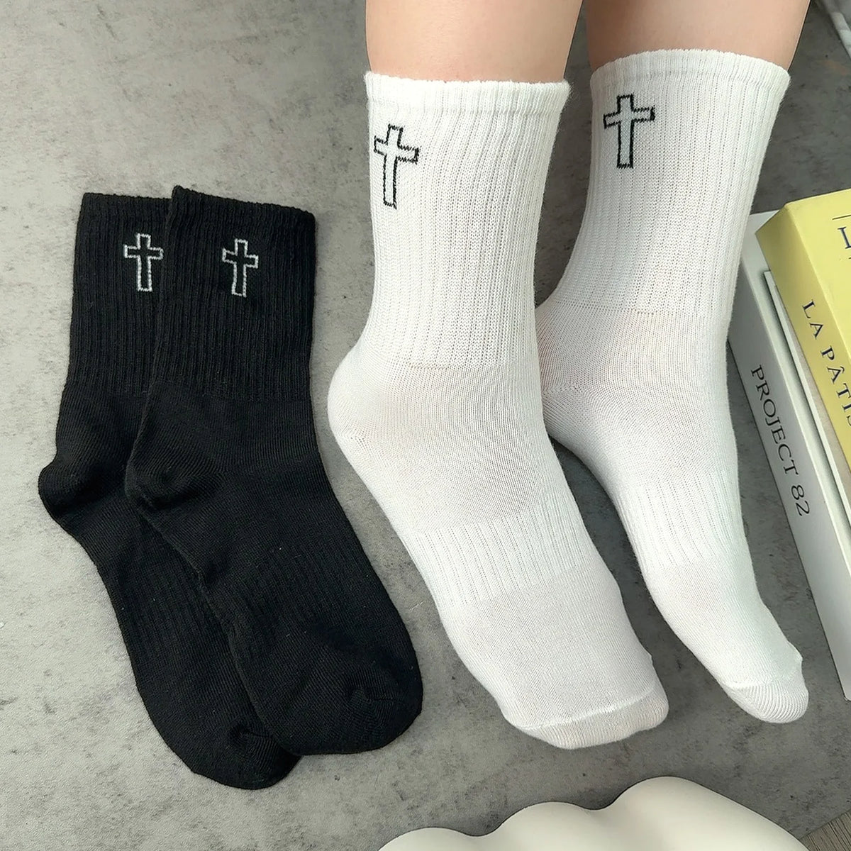 A pair of white crew socks with black crosses and a pair of black crew socks with silver crosses are displayed on a gray surf
