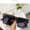 A hand holds a pair of black, oversized square sunglasses against a white background.