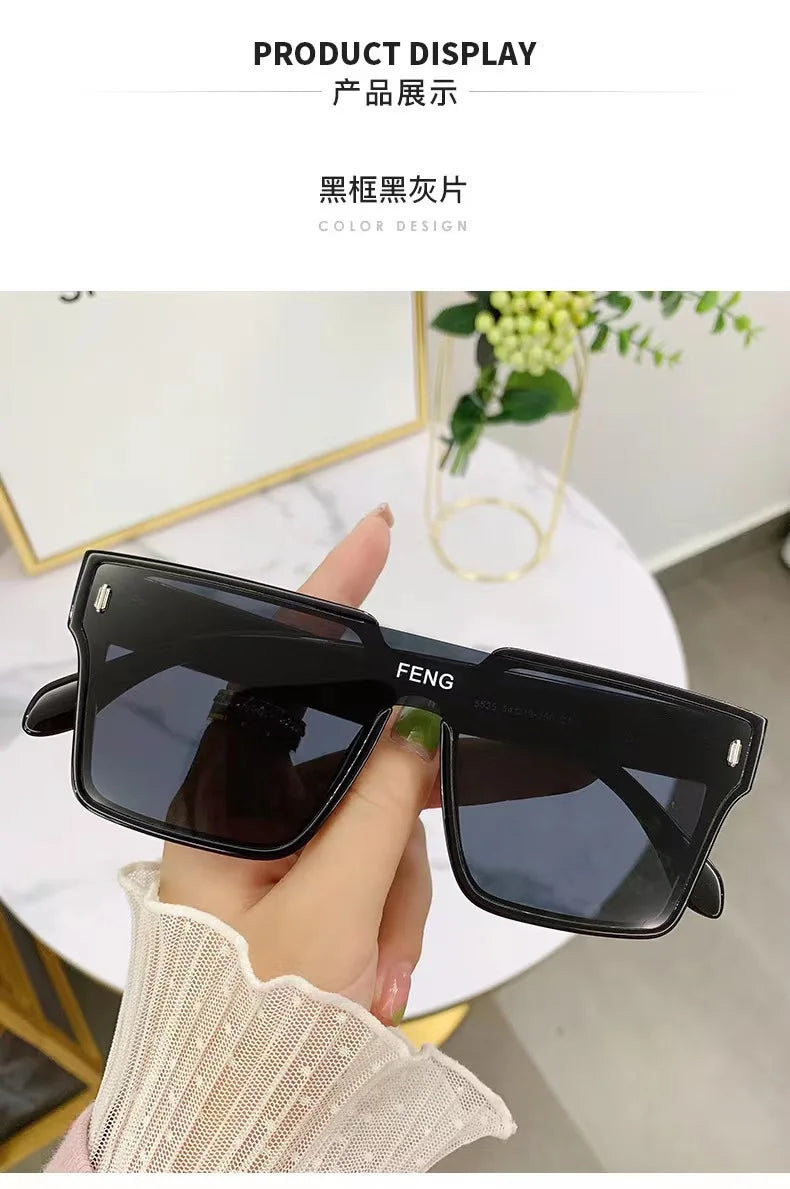 A hand holds a pair of black, oversized square sunglasses against a white background.