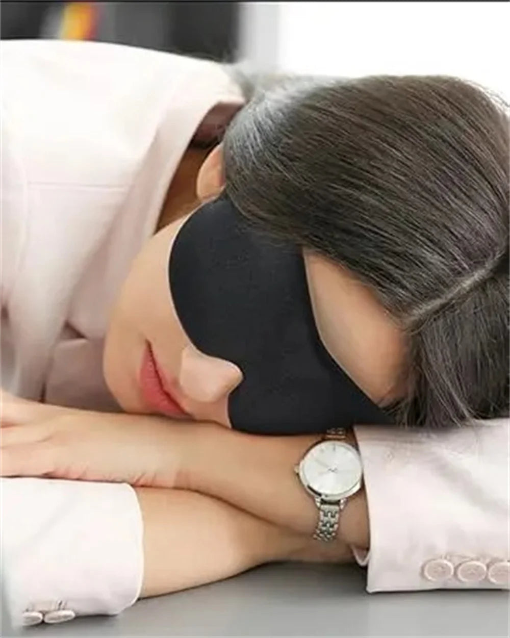 A woman is wearing a black light blocking sleep mask while resting her eyes, with a pink garment visible.