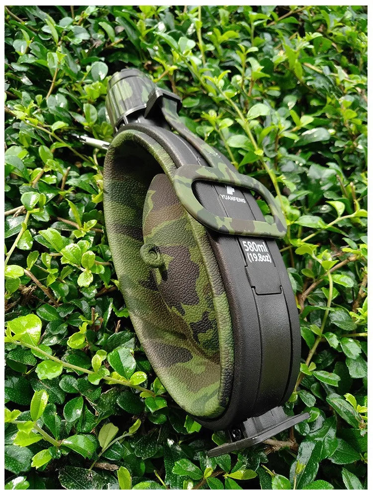 A black collapsible silicone camping kettle with a camouflage green sleeve is shown outdoors among green foliage.