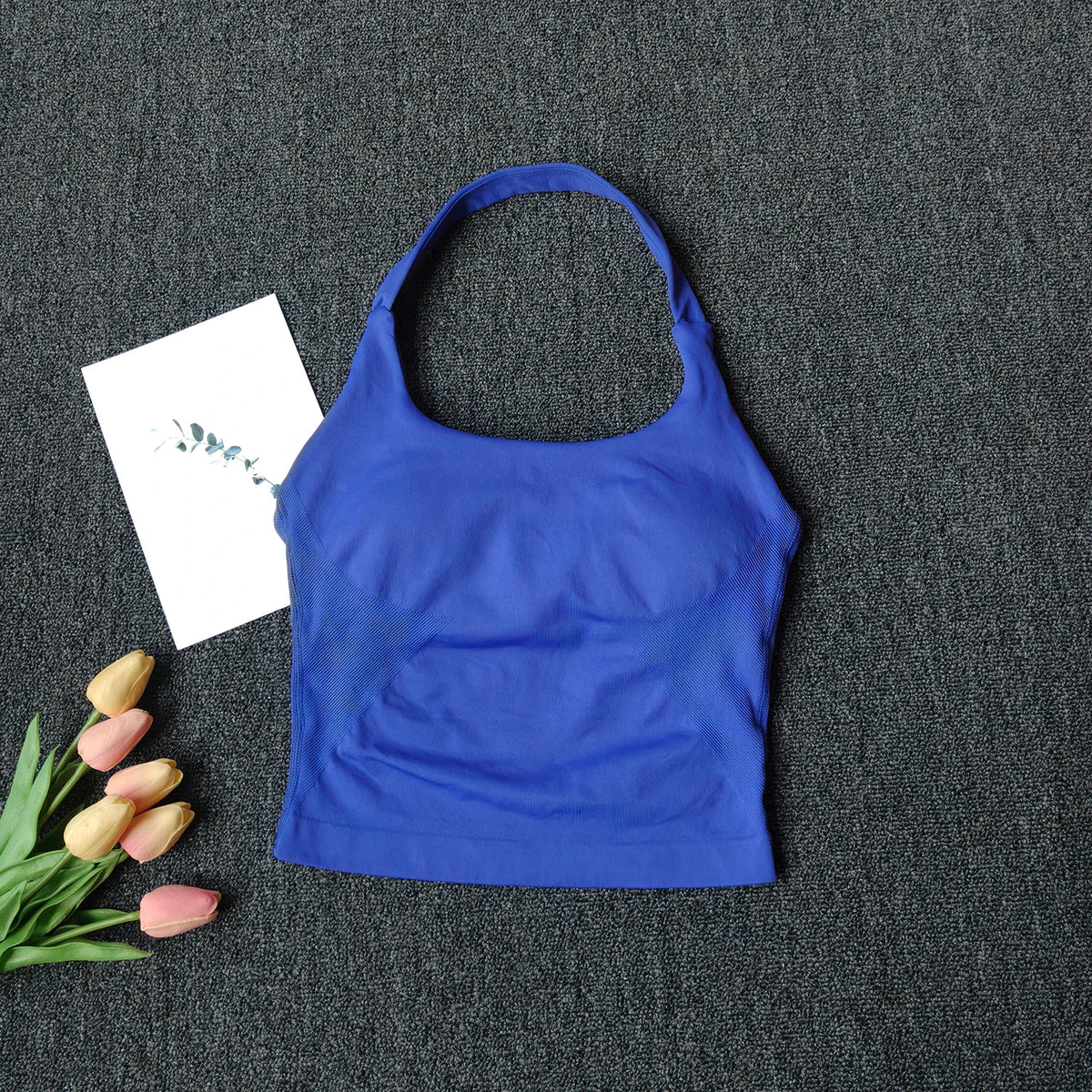 A royal blue seamless yoga and pilates sports bra laid on a dark gray fabric with pink tulips and a white card.