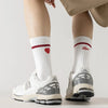 A pair of white crew socks with a red heart detail are worn with light beige pants and white athletic shoes.