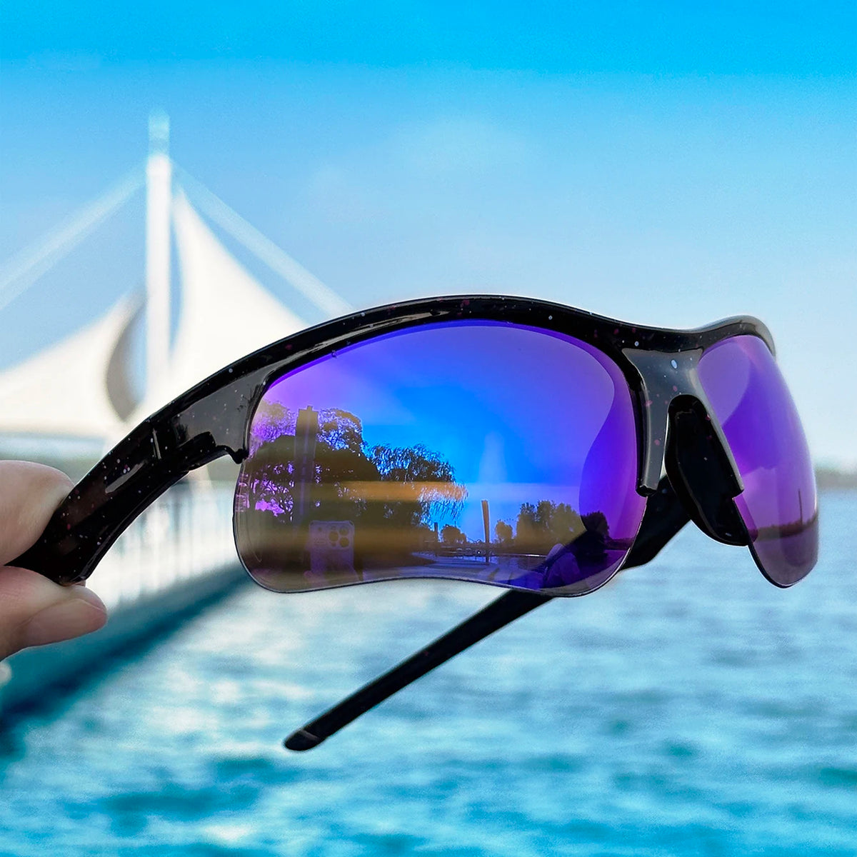 A person holds a pair of purple polarized sports sunglasses against a bright blue sky and water background.