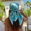 A woman ties a blue silk floral scarf around her hair outdoors.