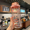A hand holds a clear pink water bottle with an orange lid and a visible time scale markings.