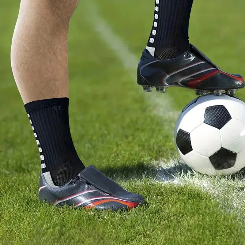 Primer plano del pie de un jugador de fútbol con zapatillas deportivas antideslizantes y calcetines negros junto a un balón.