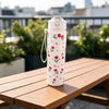 A white reusable strawberry bow water bottle standing on a wooden table outdoors.