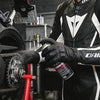 A person wearing black leather riding gloves holds a can of lubricant near a motorcycle engine.