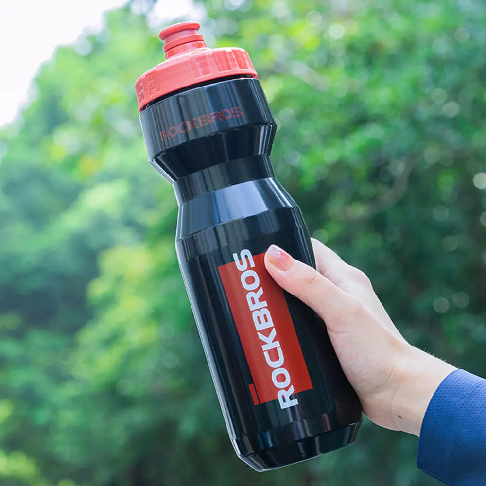 A hand holds a black sports water bottle with an orange cap against a blurred green outdoor background.
