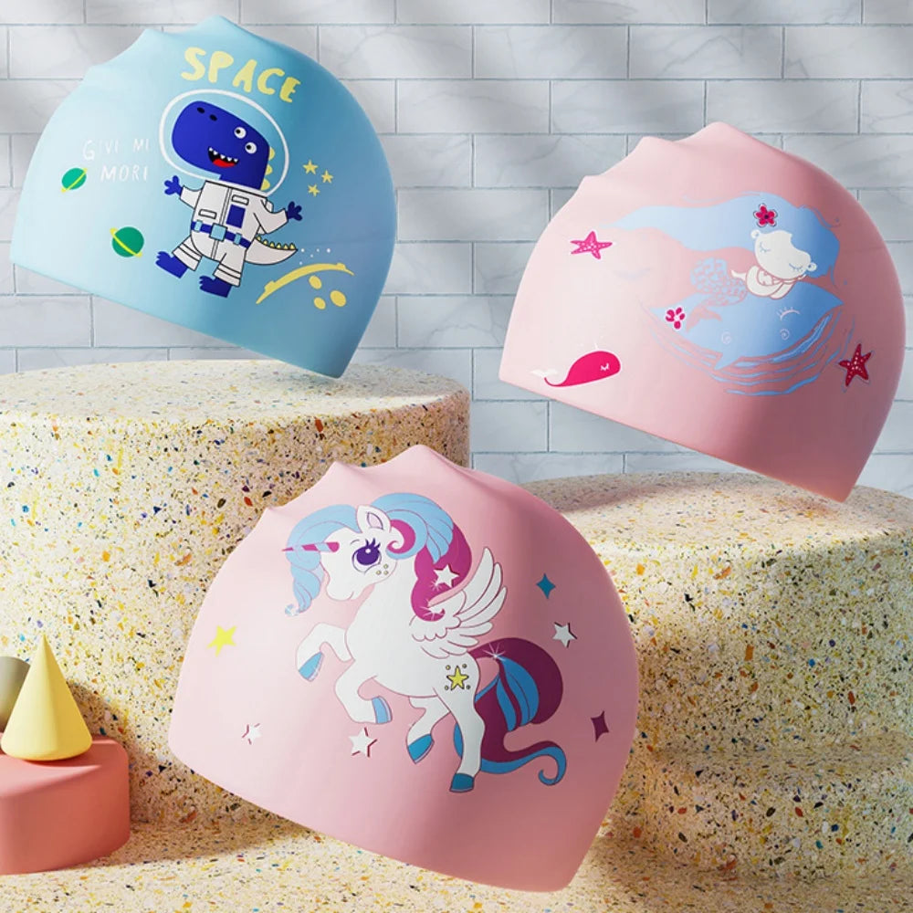 Three silicone swimming caps featuring space, a unicorn, and a dinosaur design displayed on textured surfaces.
