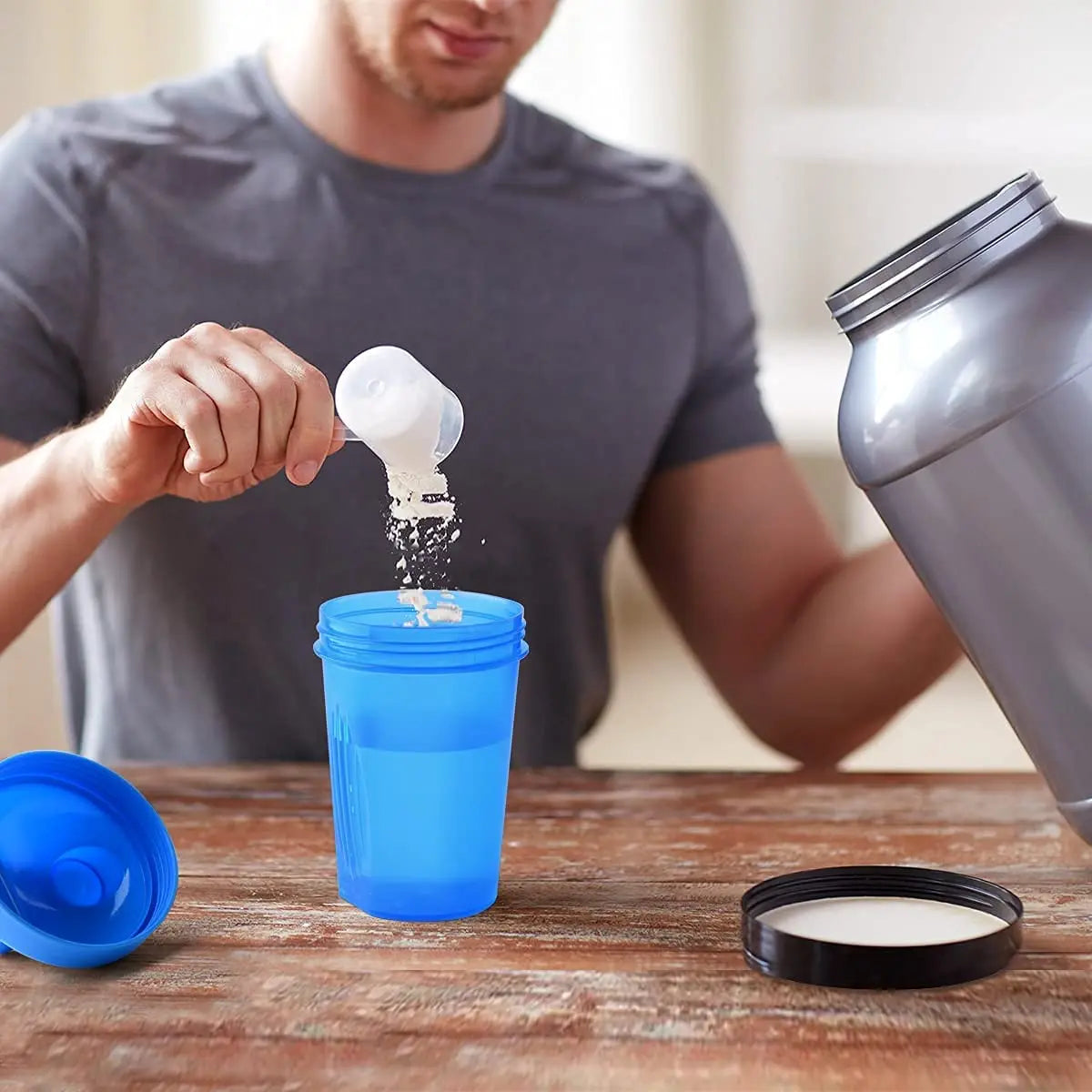 A man is pouring a white powder into a blue sports shaker bottle on a wooden table.