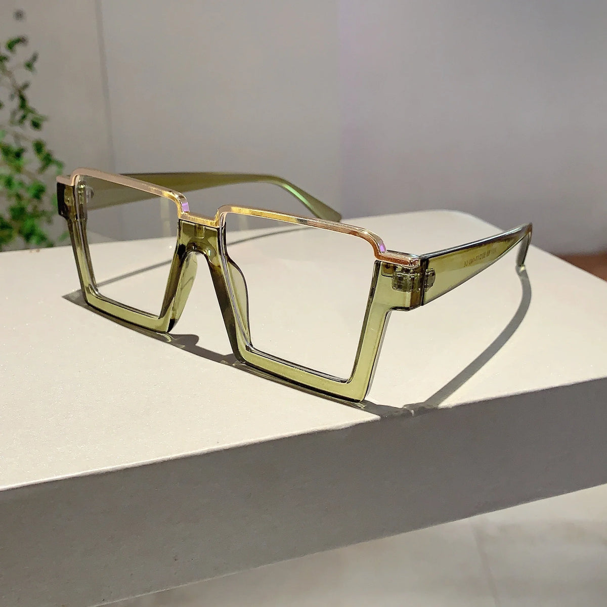 A pair of oversized square semi-rim metal eyeglasses with a light yellow or gold frame resting on a white surface.