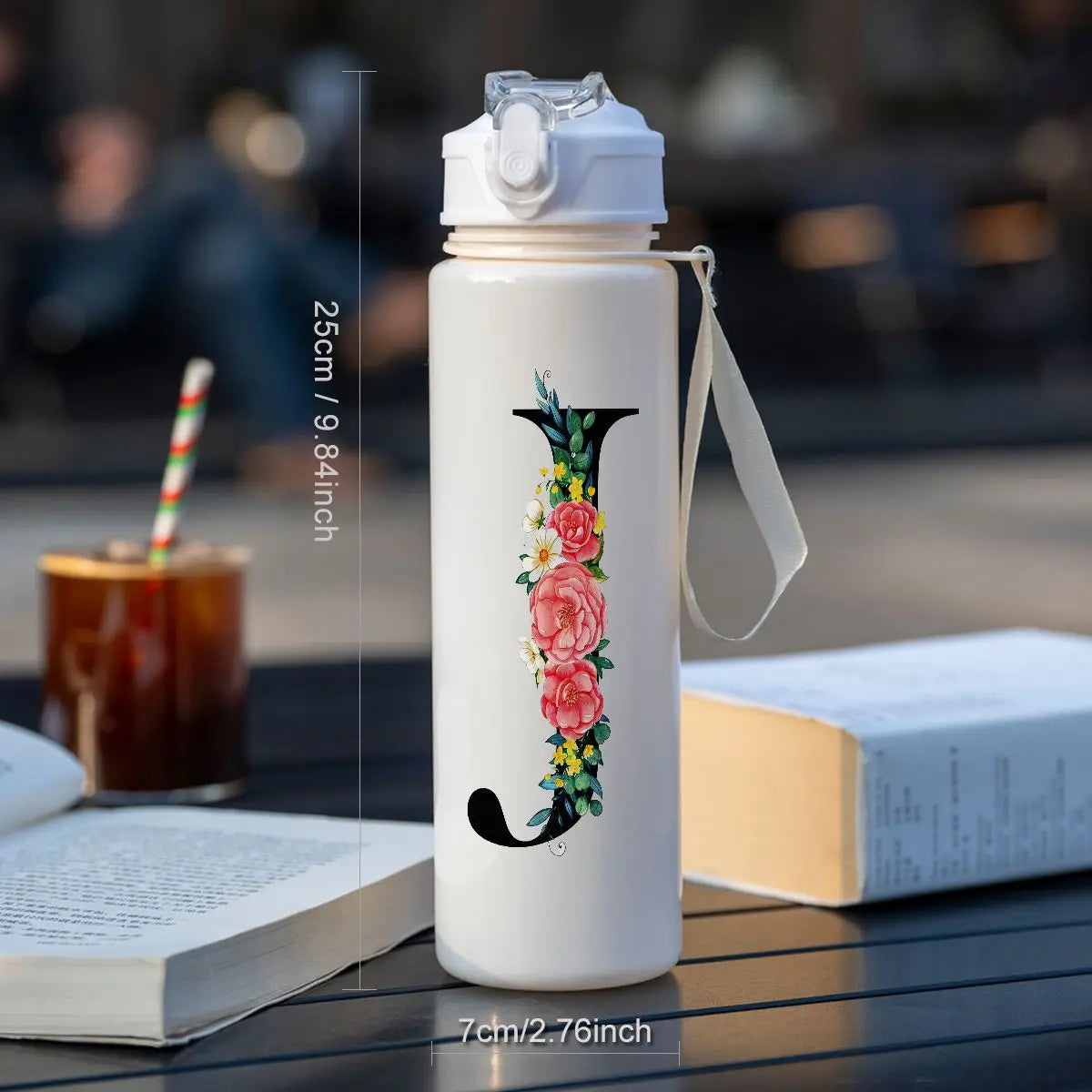 A white insulated straw tumbler with a black hook design is shown on a dark surface next to a book and a glass.