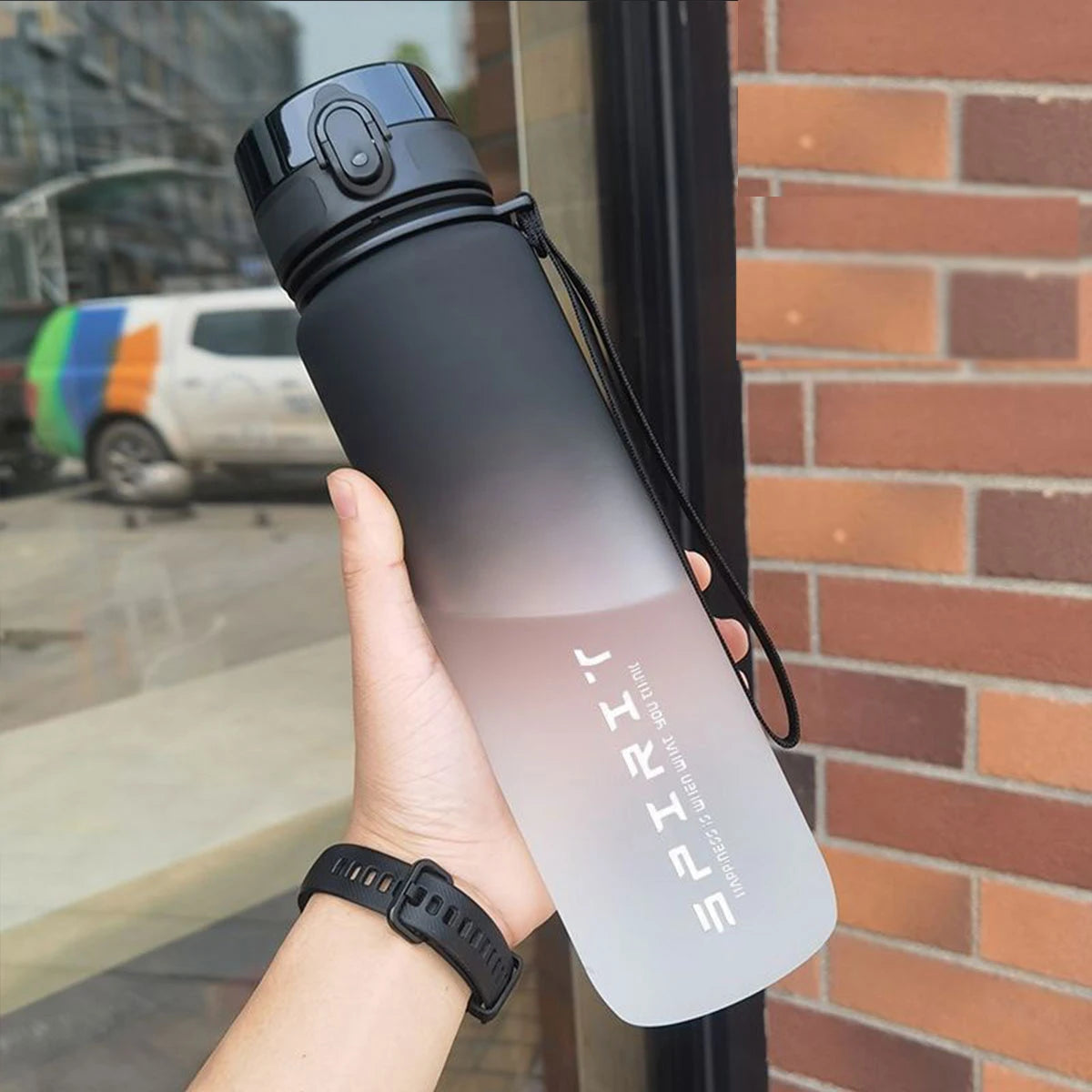 Eine Hand hält eine vergraufarbene isolierte Wasserflasche mit schwarzem Deckel vor einer Ziegelwand.