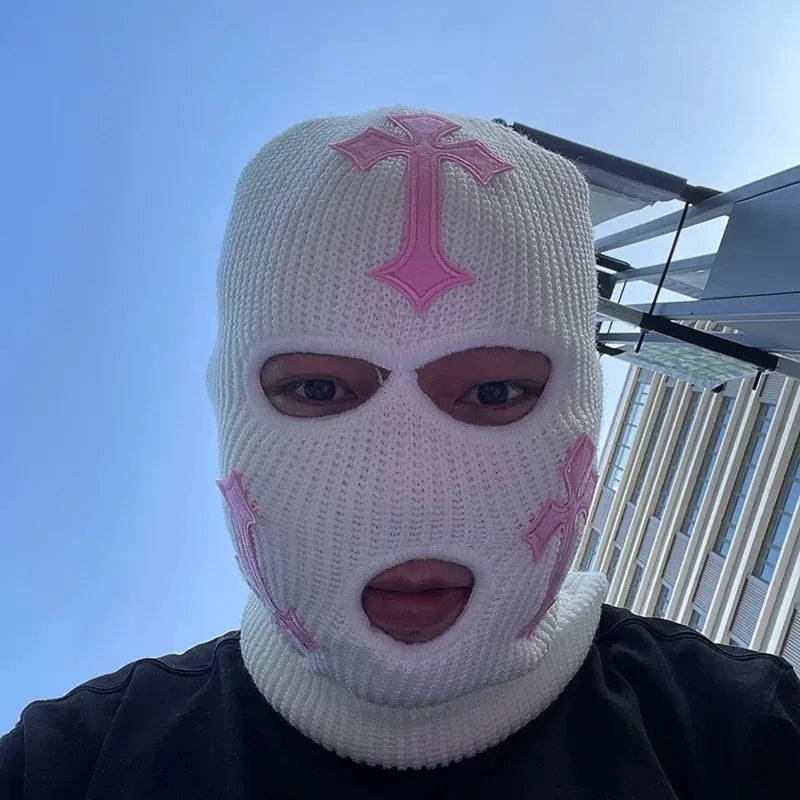 A person wearing a white knit balaclava with pink cross designs against a blue sky backdrop.