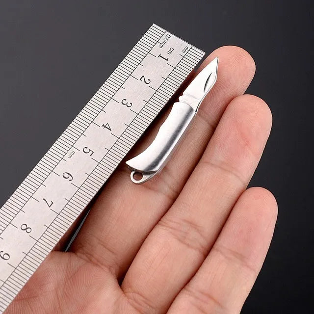 Un mini cuchillo llavero plegable de latón se muestra apoyado en una mano junto a una regla para escala.