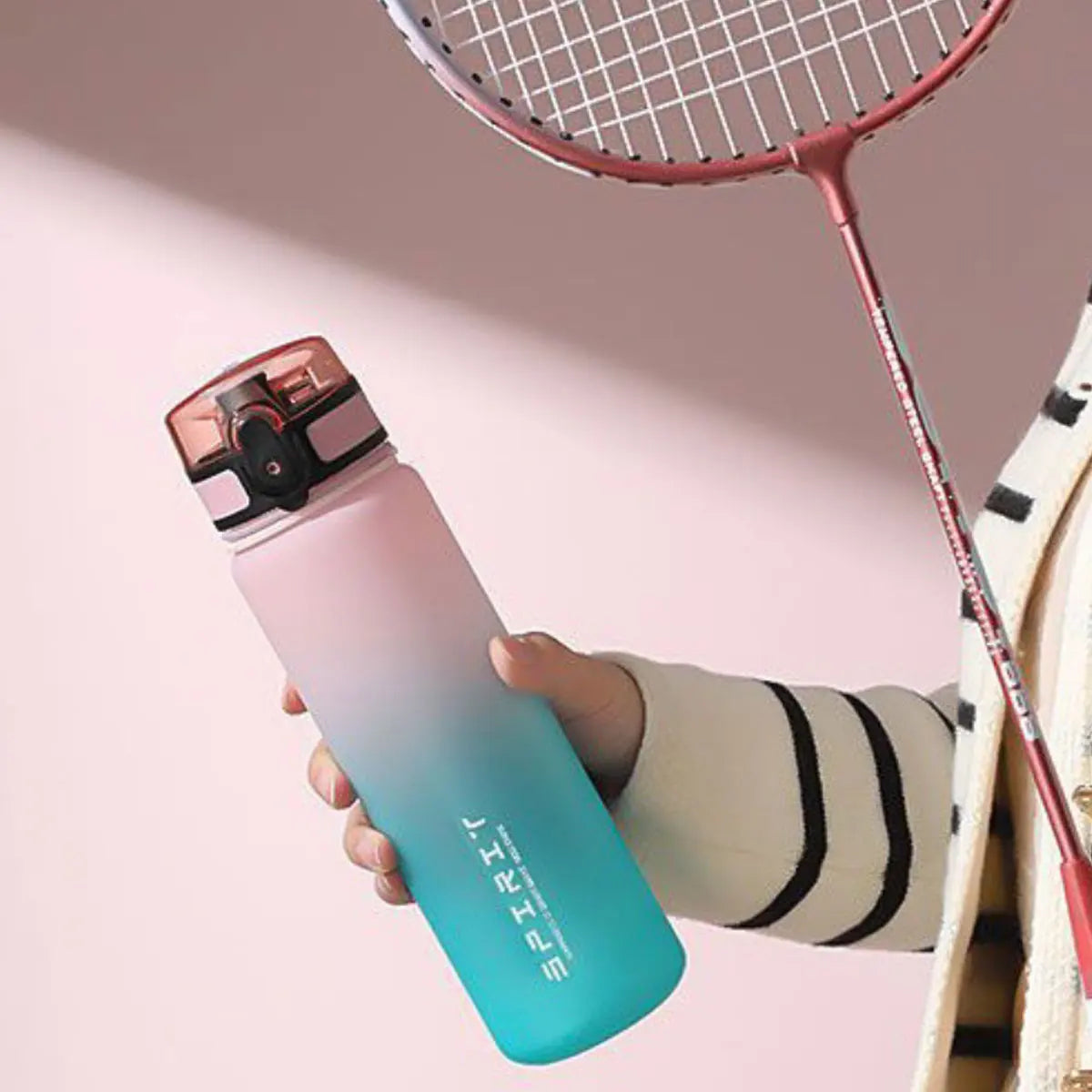 Eine Hand hält eine gradientierte isolierte Wasserflasche in rosa und blauen Tönen neben einem Tennisschläger.