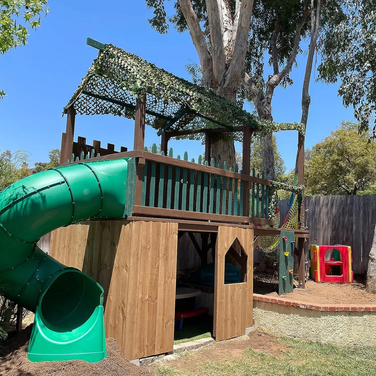 A wooden play structure with a green shade net roof and a large green plastic slide.