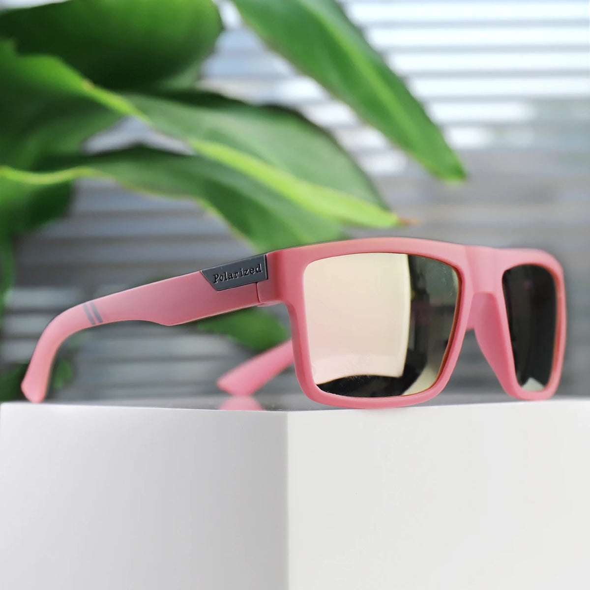 Pink polarized square sunglasses resting on a white surface with green foliage in the background.