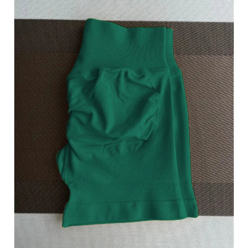 Un short de yoga sans couture, vert foncé et taille haute, est posé à plat sur une surface marron texturée.