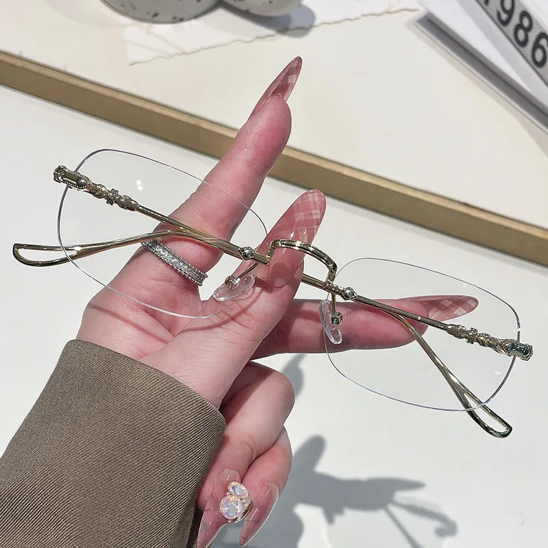 A hand holds a pair of thin, gold-toned frameless rectangle glasses with clear lenses.