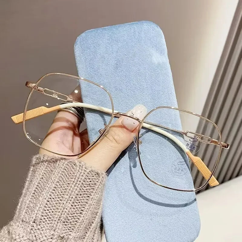 A hand holds a pair of thin, gold-rimmed reading glasses with light wood accents against a light blue background.