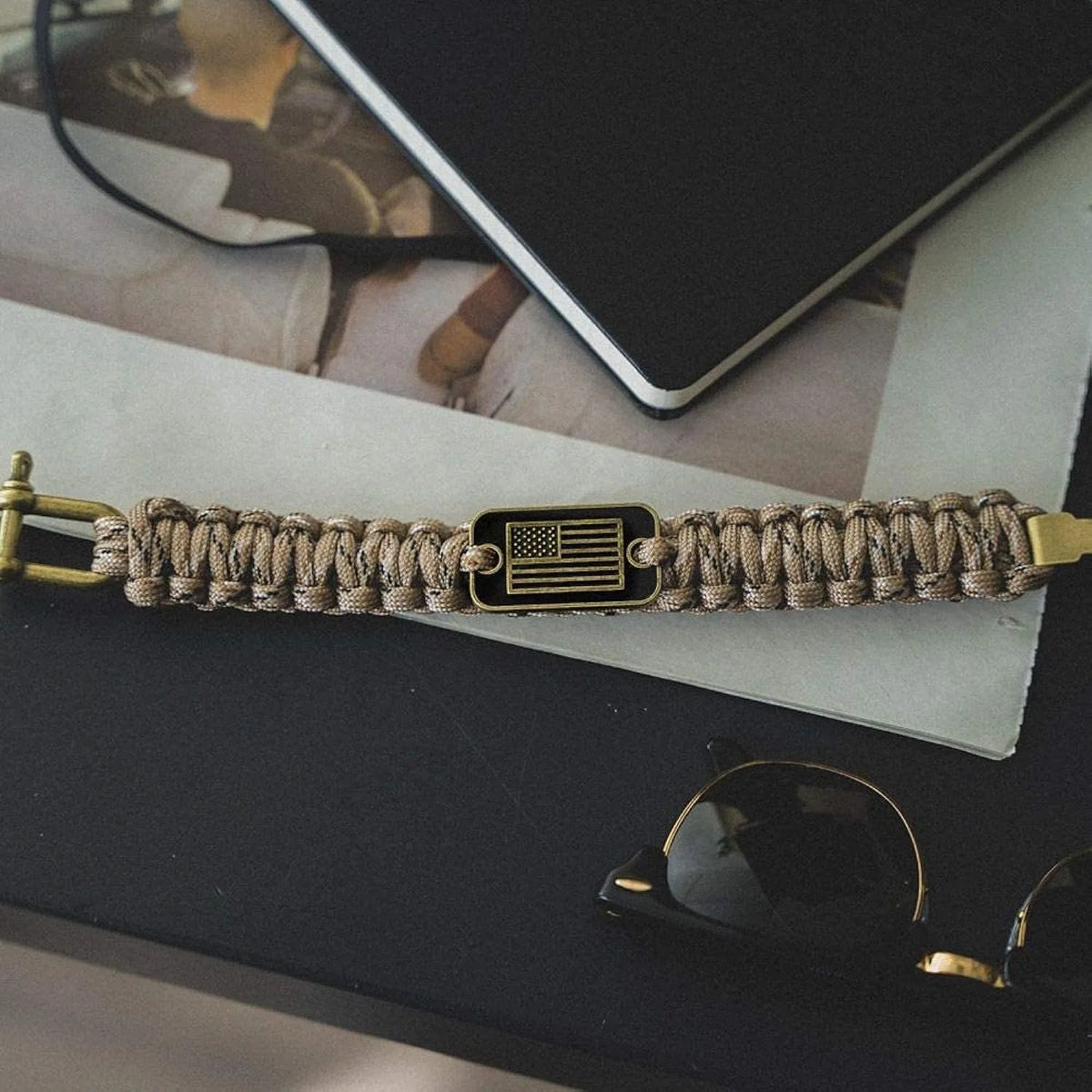 A tactical paracord survival bracelet with a bronze American flag charm resting on a dark surface.