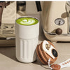 A white insulated thermos bottle filled with green latte art sits next to a brown lid and a coffee filter.