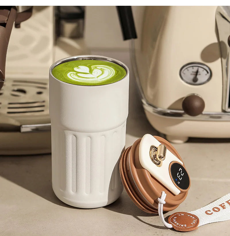 A white insulated thermos bottle filled with green latte art sits next to a brown lid and a coffee filter.