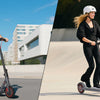 Two people ride high-performance electric scooters outdoors on a paved path.
