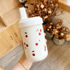 Un tumbler isotherme blanc avec un couvercle à paille décoré de petites images de cerises rouges, tenu par une main dans un m