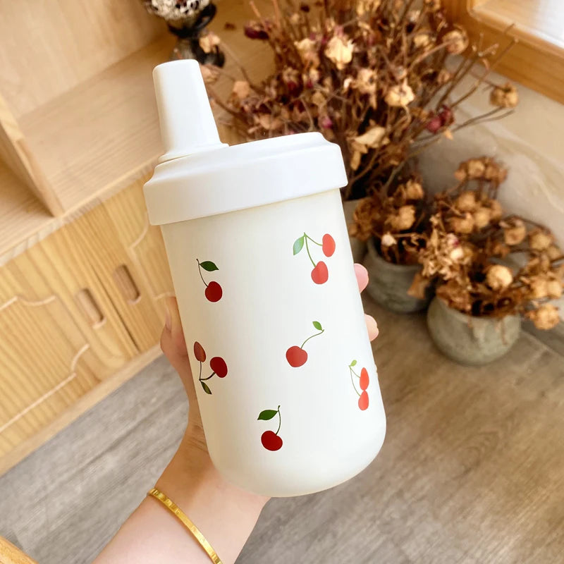 Un tumbler isotherme blanc avec un couvercle à paille décoré de petites images de cerises rouges, tenu par une main dans un m