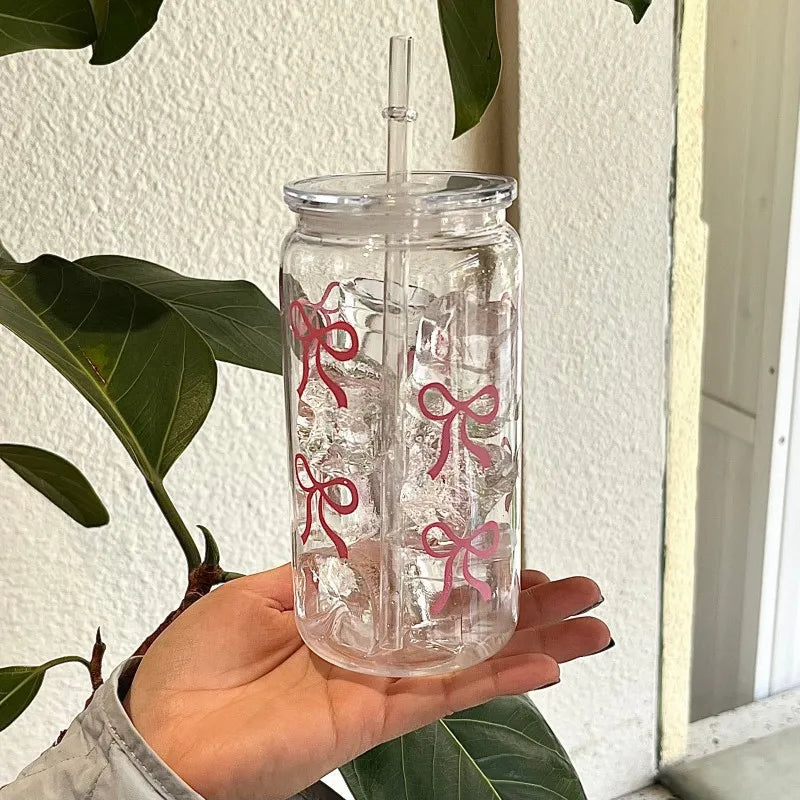 A clear glass tumbler water bottle with a pink bow design and a straw is held in a hand against a white background.