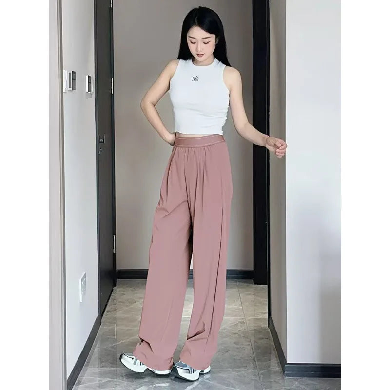 A woman models loose rose pink yoga pants and a white sleeveless top in a hallway.